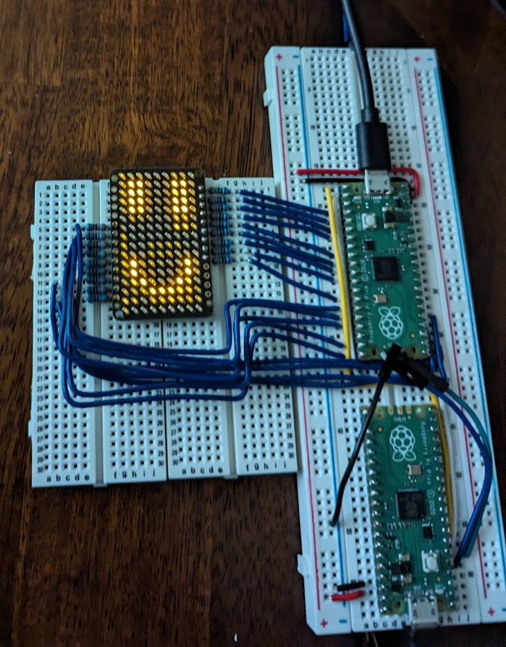 A LED grid on a breadboard with a smiley face pattern illuminated in yellow. It&rsquo;s connected with 18 blue wires to another breadboard, which has two raspberry pi picos hooked up to it.
